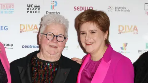 Val McDermid and Nicola Sturgeon at an event - Val McDermid has white hair, glasses, a black jacket and glittery multi coloured top. Nicola Sturgeon has brunette hair and is wearing an all pink outfit,