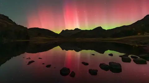 Mark Hetherington A lake in the foreground with pink and yellow light reflected in the water then the cliffs stretching away and the whole sky full of pink and yellow 