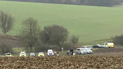 A large green field rising up a hillside, with farmland borders around the edge. Below the field, on another field, are several police cars and other emergency vehicles. 