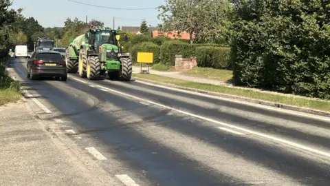 Richard Daniel/BBC A road where there are dark patches where the tarmac is melting. A black car can be seen moving away from the camera while there is a tractor coming toward the camera. 