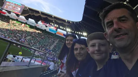 Family Amelia, Kelly, Ethan and Kevin, line up for a selfie in seats at the stadium. Crowds of fans can be seen in the stands, with part of the football pitch on view.