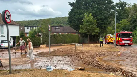 A man stands with a shovel with a woman surveying a residential street covered in mud and stones with a police officer in the background and two fire engines