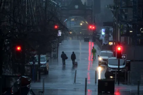 Getty Images A dark, wet day in Glasgow city centre with people walking with their jacket hoods up over their heads and street lights reflected in the wet pavement.