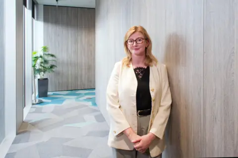 Laurence Cawley/BBC Karina Hutchins has her hands clasped in front of her in a light-filled corridor at UK Finance's HQ building in central London. Behind her is potted palm-type plant. Karina wears glasses and has stawberry blonde hair. She is smiling. She is wearing a cream-coloured business jacket and trousers and a black blouse beneath.