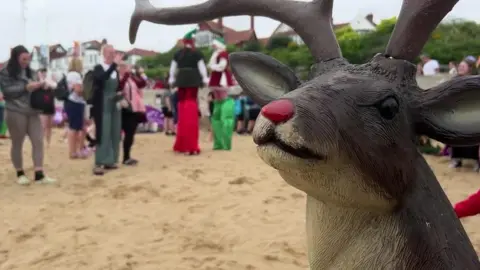 BBC A life-sized model reindeer with a red nose stands on a beach with golden sand while stilt-walkers dressed as elves walk among spectators in the background, which is out of focus.