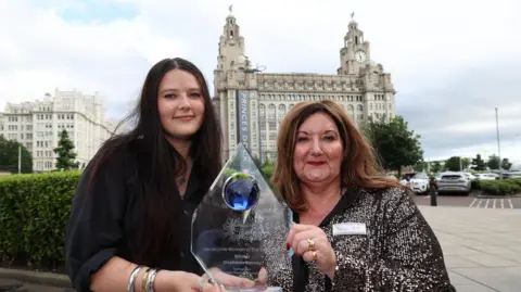 Ray Farley Stephanie Barney is presented with her trophy by Ellie Kerr in front of the Liver Building in Liverpool city centre. Stephanie has long dark brown hair and is wearing a black dress. Ellie has lighter brown long hair and is wearing a sparkly black and silver dress.