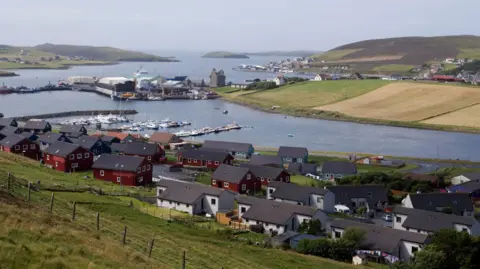 Scalloway in Shetland. The sky is blue and there are houses close to a stretch of water. There's a small ruined castle next to a harbour and green field in the background.