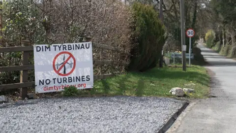 A roadside scene in a rural area, near the proposed Glyn Cothi development, showing a bilingual sign reading “DIM TYRBINAU / NO TURBINES” attached to a wooden fence beside a narrow country road, with trees, hedges and a 30mph speed limit sign visible in the background.