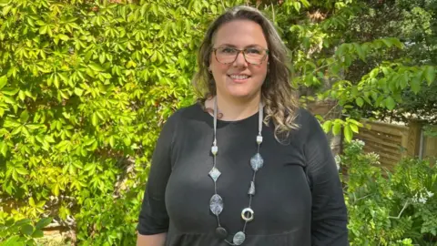 Anna Robertson has light brown hair just over shoulder length and is wearing glasses. She wears a very long necklace with different shaped rocks and rings over a black, long-sleeved top. Behind her, green leafy climbers cover a brick wall.