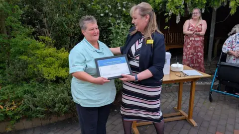 Lisa Wheelhouse has short grey hair and is wearing a light blue shirt and black trousers. She is being handed an award by another woman who has blonde hair and is wearing a pink, black and white stripy dress with a black cropped blazer