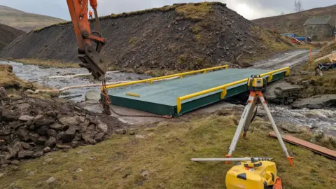 Tees River Trust A green steel structure laid across a narrow beck. The head of a crane or digger with dangling chains can be seen to one side of it and there is a large bank of earth to the other. In the foreground there is what appears to be a surveyor's instrument and a yellow tool case.