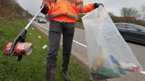 Getty Images A man in grey trousers and an orange, high-vis jacket picks up a discarded red drinks can with a litter picker. He is stood on the grass verge at the side of a dual carriageway and has a translucent bag containing more rubbish in his other hand.