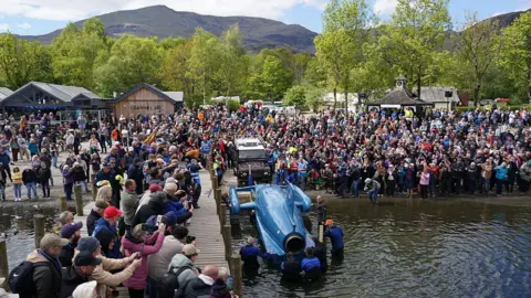 Ian Forsyth/Getty Images A team of people wearing diving equipment manoeuvre the hydroplane in the water with the help of a tow truck that brought it down the slipway. Thousands of spectators are crowded on the shore and a nearby wooden jetty. Coniston's hills can be seen in the background.