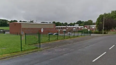 Google Red brick school buildings pictured from a distance. A road and green, metal fencing is in the foreground.