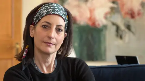 Donna looks at the camera as she sits on a blue sofa. She has a black jumper on and a head scarf with floral patterns on it. She has dark brown hair which comes down to her shoulders. Behind her on the wall is a painted flower pot with pink flowers inside.