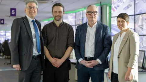 The four are stood in an office and are all smiling at the camera. Mr Foster and Mr Parker are both wearing a jacket shirt and trousers and Mr MacDonald has a grey shirt on and black trousers. Ms Marston has a light beige jacket on with a white shirt and dark trousers.