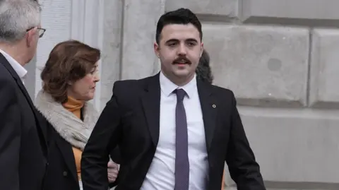 PA Media A man with black hair and a moustache, wearing a black suit and white shirt. There is a man and a woman standing behind him. 