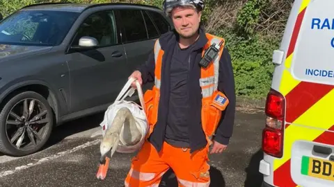 A man wearing orange high-vis uniform and a hard hat while holding an injured swan inside a specialist animal bag