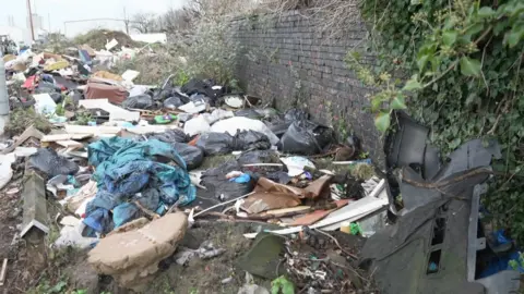 Image of a rubbish. Lots of waste, bin bags, wooden panels and pieces of cloth are strewn on the floor.
