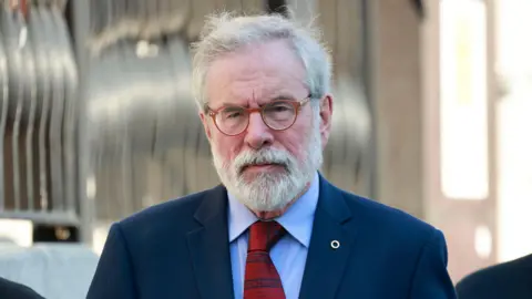 Gerry Adams walks outside the High Court in Dublin. He has short white hair and beard. He is wearing small, orange circular glasses and his expression is neutral. He is also wearing a navy suit jacket, a light blue collared shirt and a red patterned tie. On his suit jacket lapel he is wearing a silver fáinne, denoting his Irish speaking ability.