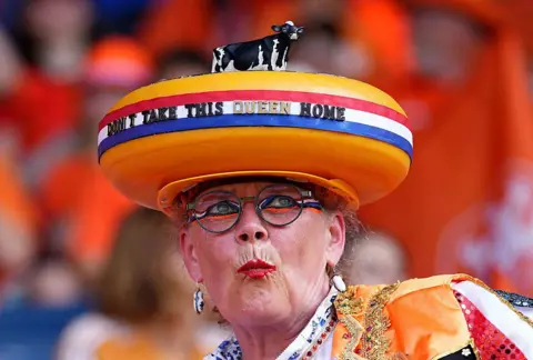 Getty Images A middle-aged woman wears a hat resembling a Gouda cheese. The hat has a striped ribbon in the colours of the Netherlands flag displaying the words "Don't take this queen home". She is also wearing red lipstick and red, white, blue and orange eye make-up