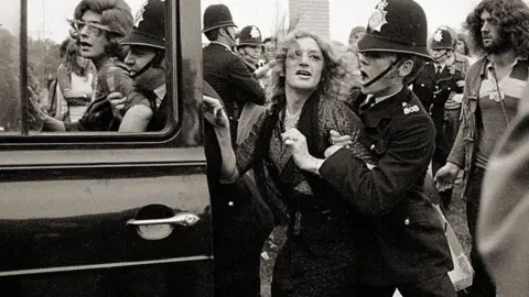 Alamy/Mirrorpix a drag queen is bundled into a 1970s police car