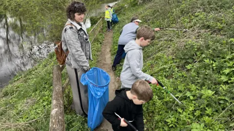 A group of young boys are using litter grabbers on a bank to clear plastic away from the river behind them. A woman is holding a large blue bag to collect litter. Other volunteers in hi-vis jackets can be seen further up the path with another blue collecting bag.