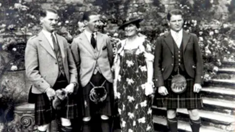 Black and white photo of three men in kilts, either side of a lady - their mother - in a floral dress.