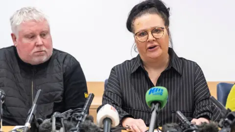 PA Media A man with white hair and stubble sits next to a woman with dark hair and glasses at a press conference. They are sat behind a desk. Radio microphones are placed on the desk and pointed in their direction.