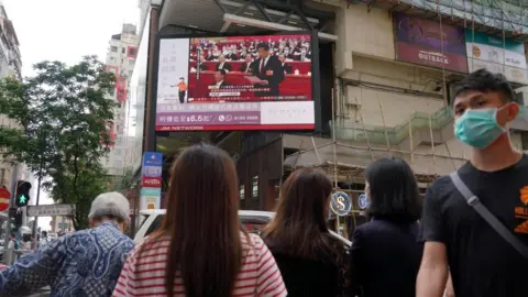 Getty Images People wearing face masks walk past a large screen in Hong Kong displaying a live broadcast in the street of the National Congress