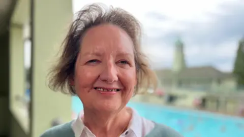 Emma Baugh/BBC Jane Callaghan is standing on a balcony which looks over Peterborough Lido. Callaghan has short brown hair and is wearing a white shirt and green jumper.