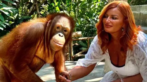 Kelly Kershaw is kneeling down in a white dress holding the hand of an orangutan, which is looking at the camera. Behind them is a forest environment.