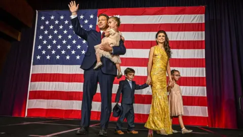 Getty Images Ron DeSantis, his wife, Casey, and their three children pose in front of a United States flag. Mr Santis, wearing a blue suit with a white shirt and red tie, holds one of his young daughters, waving with his free arm. Mrs Desantis, wearing a fitted gold/yellow floor-length dress, stands with the couple's young son and daughter at her side, holding their hands.