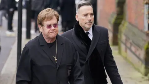 PA Media Sir Elton John and his husband David Furnish attending the funeral service of Derek Draper