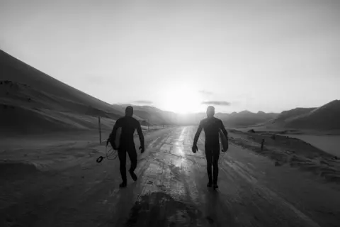 Thomas Meurot Surfers wearing wetsuits and holding surfboards walk along a path in Iceland