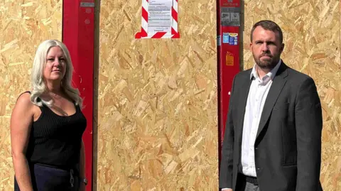 Newcastle-under-Lyme Borough Council Jonathan Gullis standing next to Venessa Renshaw outside a shop which has been boarded up. A closure notice has been taped to the front of the store.