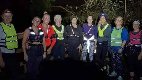 Mandi Burroughs The image shows a group of women standing together at night. They are all smiling at the camera, wearing hi-vi clothing.