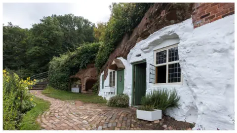 A charming white cottage is built into a red rock cliff face. A cobblestone path leads to the entrance, with green doors and windows opening to the exterior.