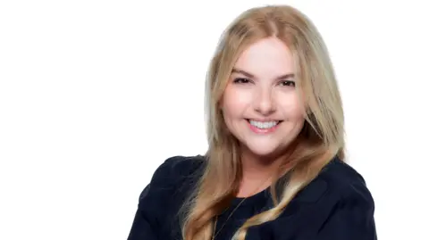 Danny Evans A head and shoulder shot of a woman with long blonde hair, wearing a black top and long gold necklace. She is looking into the camera and smiling. It's a professional photo with a white background.