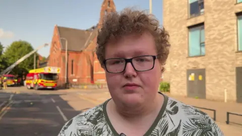 Liam Keatley, who wears glasses, stands in front of a cordon. A church and two fire engines can be seen in the background