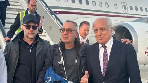 Handout via Reuters George Glezmann (centre) stands at Kabul airport before boarding a flight to Qatar. He is visibly emotional in the photo. To his right is US hostage envoy Adam Boehler and to his left is US's former envoy to Kabul, Zalmay Khalilzad. Qatari officials were also present