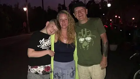 Three people standing close together in the foreground on a dimly lit street at night, one leaning head against another. Clothing includes a black T-shirt with white text, a bright green sleeveless layer, and a green printed T-shirt with a graphic face. A hand-held bag is partially visible at waist level. In the background, street lamps, parked cars, and trees line the road, with scattered lights and silhouettes visible in low light.