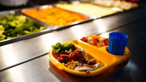 A school meal in an orange tray with vegetables and meat in one side and strawberries in the other side and a blue plastic cup. In the background are containers with food in