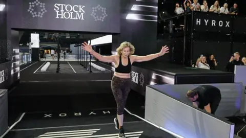 Hyrox/Annie Reid A woman crosses the finish line of a Hyrox race with her arms outstretched in celebration.