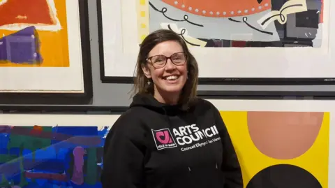 Suzanne, a woman with short brown hair, she wears glasses and has a bright smile as she stands in front of bright contemporary art.