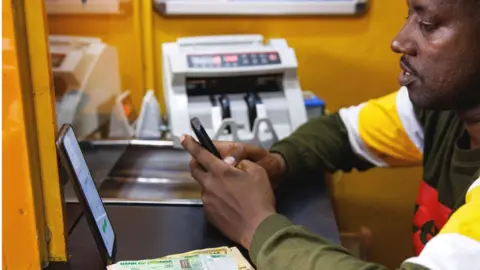 Getty Images An agent carries out a mobile money transfer transaction with a customer inside a kiosk in Kampala, Uganda, on Wednesday, Aug. 16, 2023.