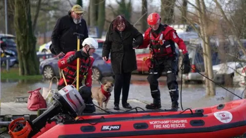 Jacob King/PA Media Rescue personnel in red clothing assist a resident in a brown coat onto a red inflatable boat