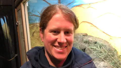 Darren Rozier/BBC Zoe Nunn with light brown hair smiles at the camera. She is wearing a blue coat with a blue top underneath. She is standing in front of an animal enclosure which has a glass window front.