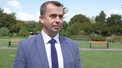 Bicester and Woodstock MP Calum Miller is wearing a blue blazer and tie and is stood in front of a grass field.