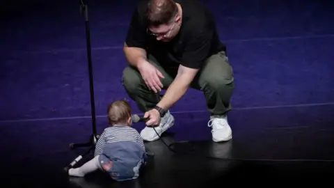 A baby on stage with a man crouching down holding a micophone. The baby is wearing a blue striped top. The man is wearing green trousers with a black tshirt and white trainers.